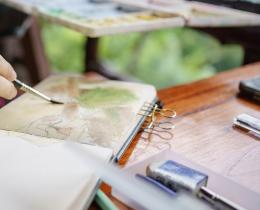 Person painting with watercolors in a journal, symbolzing a creative expression workshop or arts retreat.