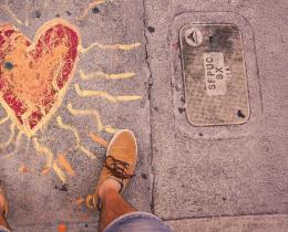 Person standing on a sidewalk with a heart and radiating sun rays painted beneath their feet, symbolizing leaning inward to love, empathy, and anchoring into one’s spiritual center.