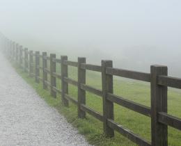 Foggy gravel path between wooden fences disappearing into mist, symbolizing liminal space, personal growth, transition, uncertainty, grief, and new beginnings