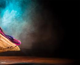 Kathak dancer in a flowing traditional costume performs on stage, surrounded by dramatic lighting and colorful smoke, symbolizing the fusion of movement, culture, and spiritual expression through dance and Ayurveda.