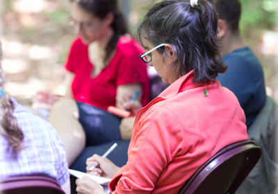 Woman in pink shirt sits in a circle with others, writing in a notebook.