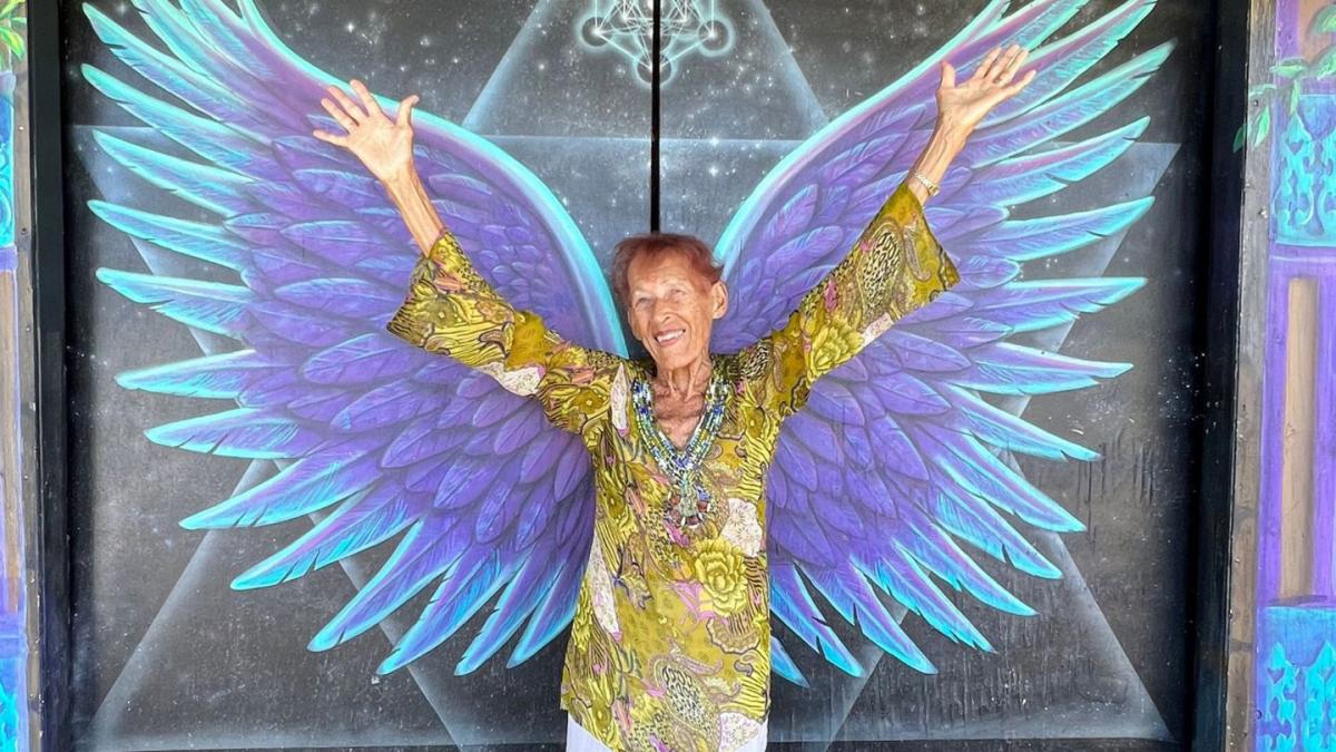 Dr. Beverley Salmon, smiling and arms raised against a backdrop with purple and blue angel wings.