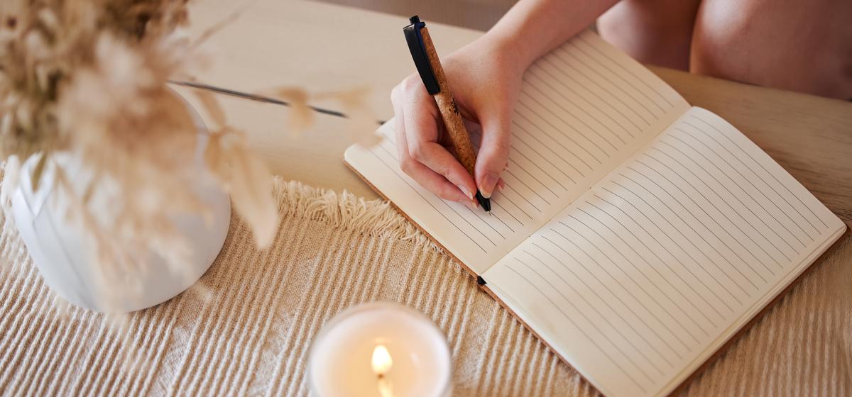 Person writing in a journal on a cozy desk with flowers and a candle