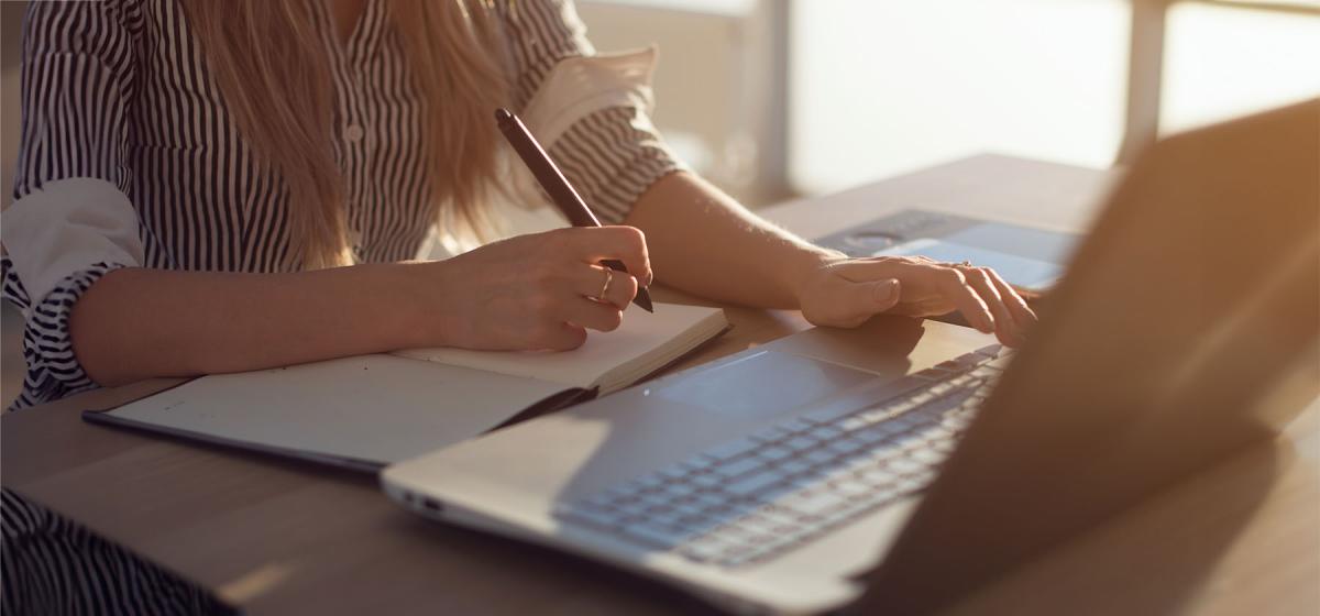 Person writing in a notebook while looking at a computer