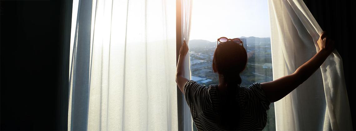 Woman opening bedroom curtains to welcome morning sunlight, symbolizing a peaceful morning routine and mindfulness ritual for starting a calm and centered day.