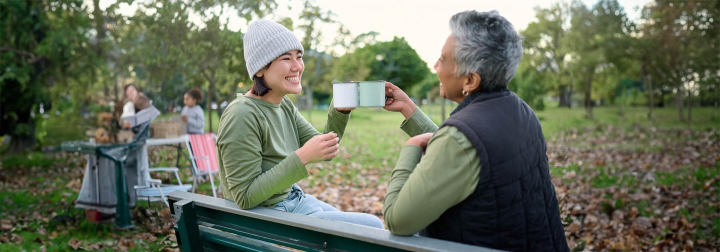 Two people smiling and clinking mugs together on a park bench, symbolizing friendship, connection, and practices that alleviate loneliness.