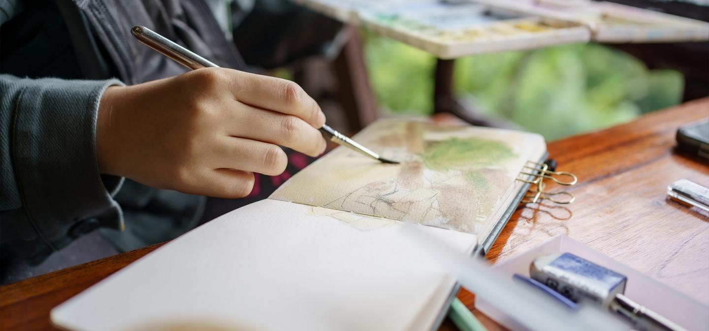 Person painting with watercolors in a journal, symbolzing a creative expression workshop or arts retreat.