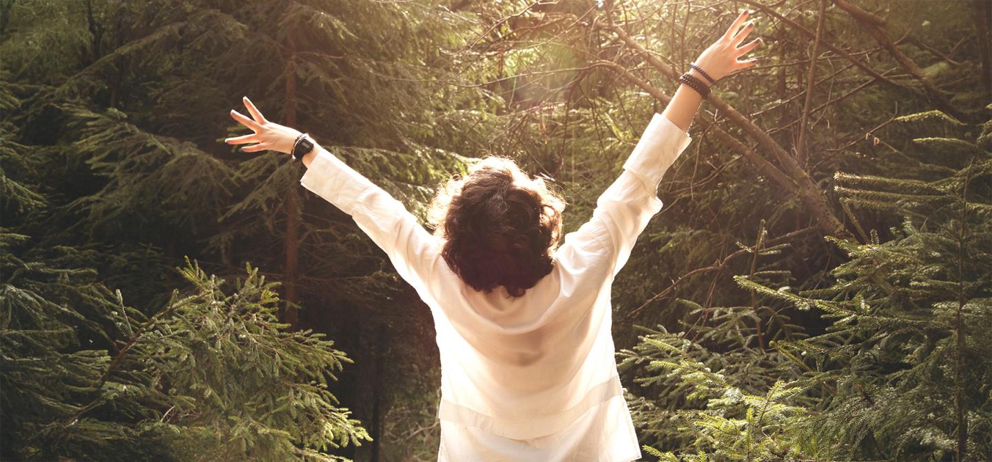 Person standing in a sunlit forest with arms raised, embracing a moment of joy and connection with nature.