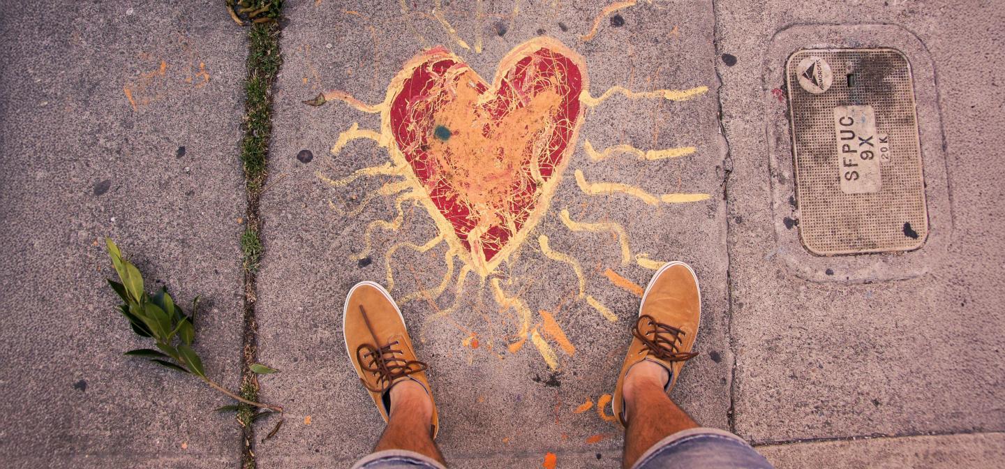Person standing on a sidewalk with a heart and radiating sun rays painted beneath their feet, symbolizing leaning inward to love, empathy, and anchoring into one’s spiritual center.