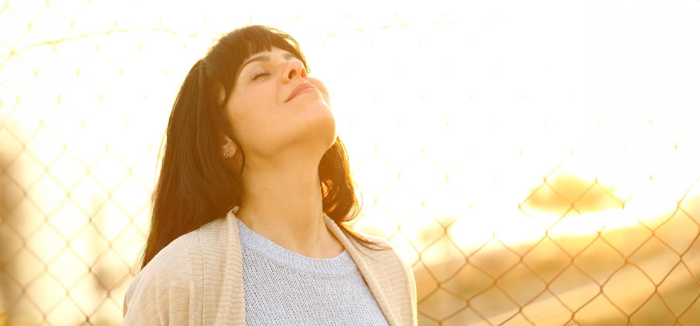 Woman standing outdoors with eyes closed in sunlight, symbolizing calm breathing and nervous system regulation through daily rhythm.