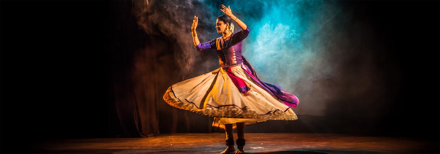 Kathak dancer in a flowing traditional costume performs on stage, surrounded by dramatic lighting and colorful smoke, symbolizing the fusion of movement, culture, and spiritual expression through dance and Ayurveda.