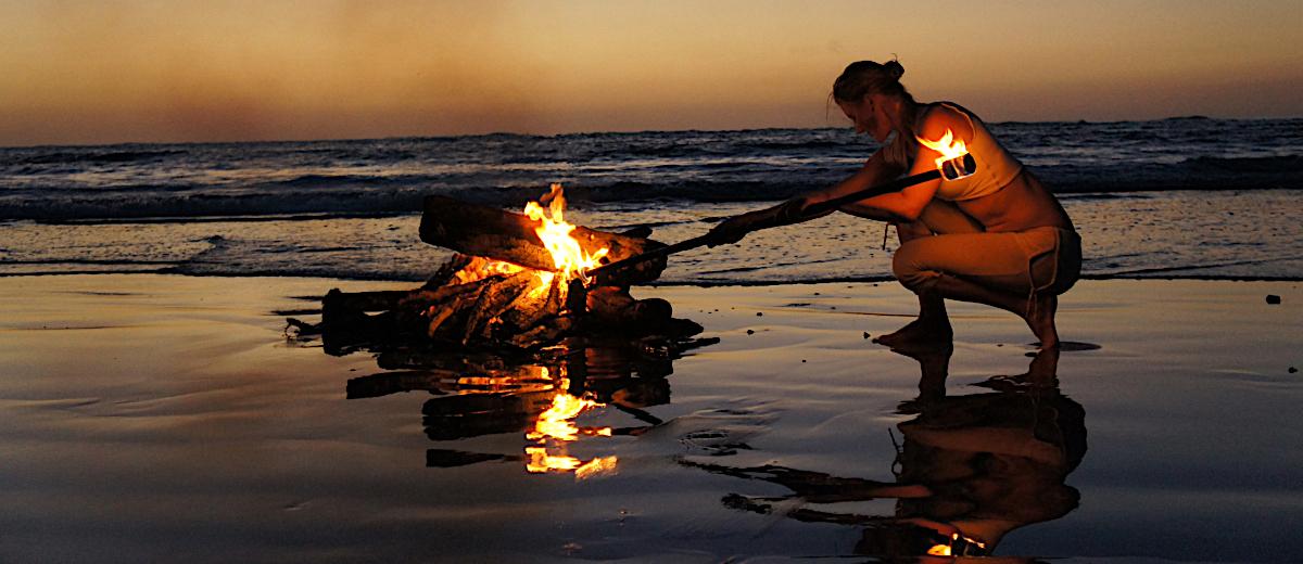 Woman tending a beach bonfire at sunset, reflecting the power of nature-based rituals and sacred traditions during a coastal retreat for regeneration and spiritual renewal.