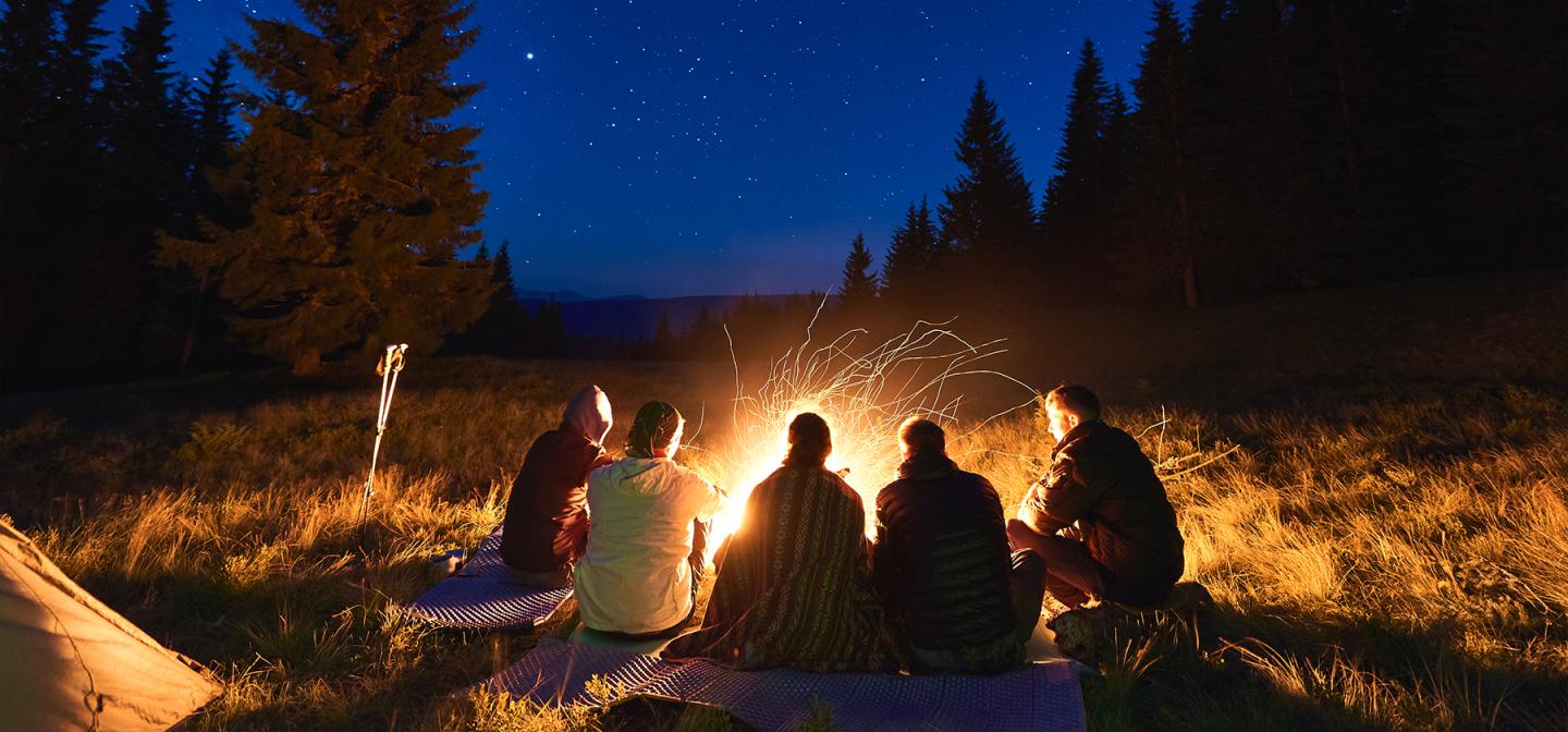 Five people sitting around a glowing campfire under a starry night sky, surrounded by trees—evoking the ancient tradition of storytelling in community.