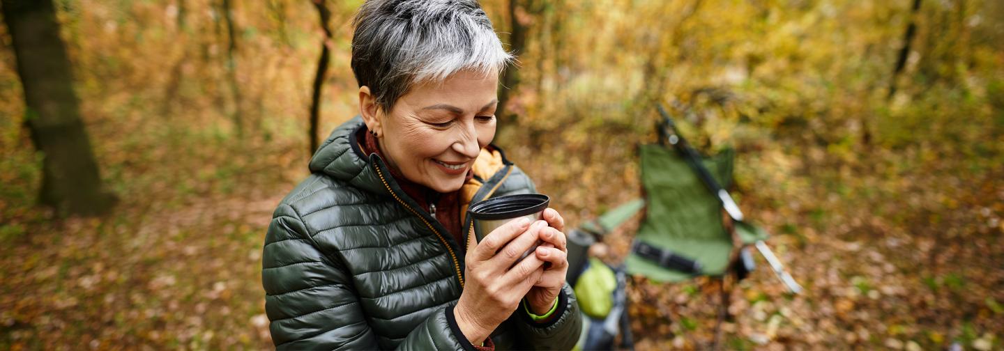 Woman enjoying a quiet moment outdoors in midlife, holding a warm drink in an autumn forest.
